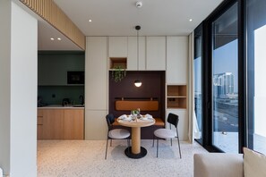 Un intérieur moderne avec une table de salle à manger ronde, deux chaises, un luminaire suspendu et une vue sur le paysage urbain à travers de grandes fenêtres.