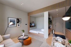 A modern hotel room with a bed, a sitting area with a sofa and a small table, a dining table set for two, and a kitchenette in the background. - Cozy Studio w/ Burj View Balcony, Business Bay (Dubai)