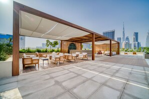 An outdoor seating area with multiple chairs and tables, a wooden pergola, and a city skyline in the background. - Cozy Studio w/ Burj View Balcony, Business Bay (Dubai)