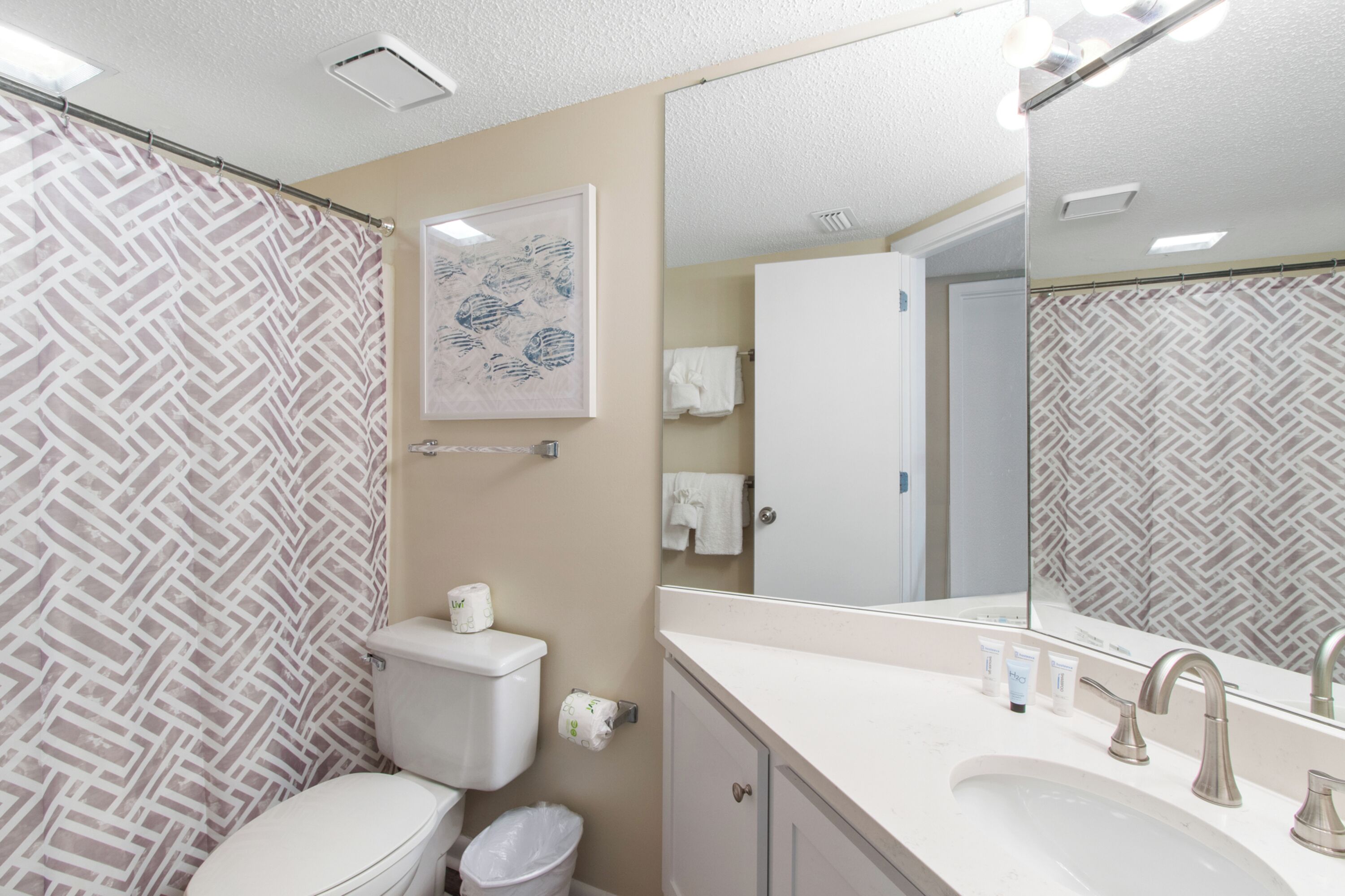 Une salle de bain avec un rideau de douche à motifs, des toilettes, un lavabo avec un miroir et un tableau encadré au mur.