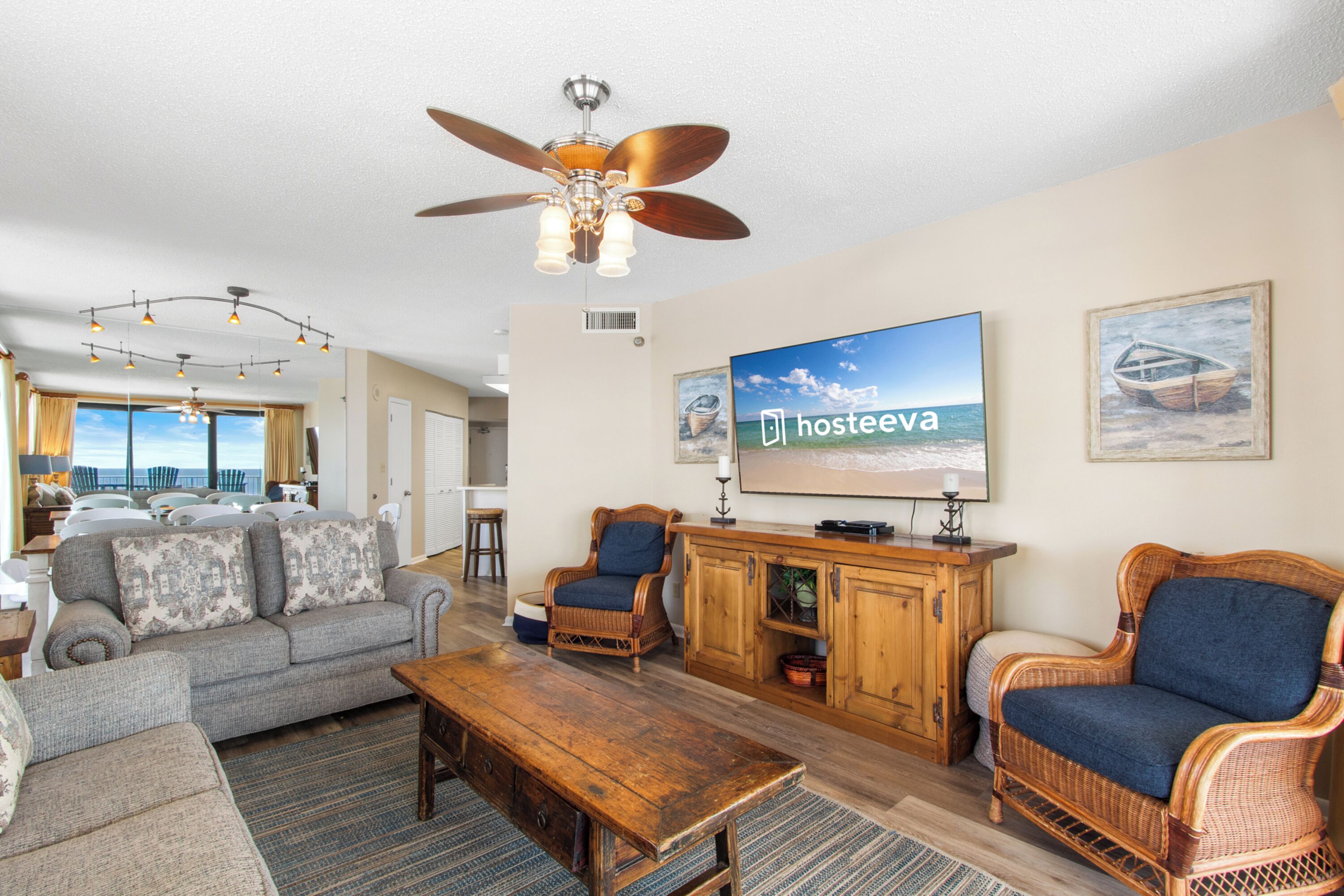 A spacious living room with a large flat-screen TV, wicker furniture, and a view of the ocean through large windows.