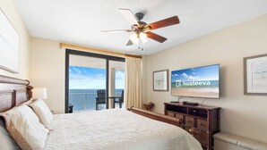 Habitación de hotel con una cama grande, ventilador de techo, televisor y vista al océano desde las puertas del balcón.