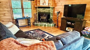 Interior de una cabaña de troncos con chimenea, televisor, sofá y ventilador de techo.