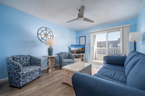 Ein Wohnzimmer mit einem blauen Sofa, zwei gemusterten Sesseln, einem kleinen Tisch mit Lampe, einem Deckenventilator und einem Fernseher auf einem Schrank.