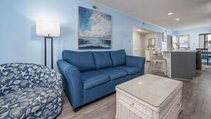 Un salon avec un canapé bleu, un fauteuil à motifs, une table basse en osier et un tableau représentant des nuages accroché au mur.