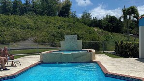 Una piscina con una fuente de piedra, rodeada por un pavimento de ladrillo y una valla metálica.