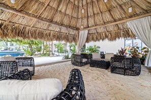 An outdoor lounge area with thatched roof, wicker furniture, and a view of a pool.