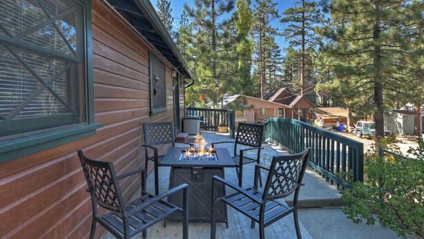 A wooden cabin with a patio featuring a fire pit and chairs, surrounded by tall pine trees. - Forest Cottage · Hot Tub · Firepit · Fenced Yard (Big Bear Lake)