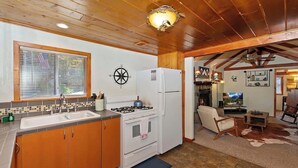A kitchen with wooden cabinets, a stove, and a refrigerator. There is a window with blinds, a compass on the wall, and a television in the living area. - Forest Cottage · Hot Tub · Firepit · Fenced Yard (Big Bear Lake)