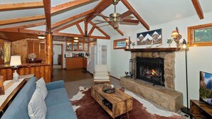 A spacious living room with a stone fireplace, a blue sofa, and a wooden ceiling structure. - Forest Cottage · Hot Tub · Firepit · Fenced Yard (Big Bear Lake)