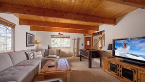 A cozy living room with a sofa, coffee table, and TV. There's a wooden ceiling and a staircase leading upstairs.