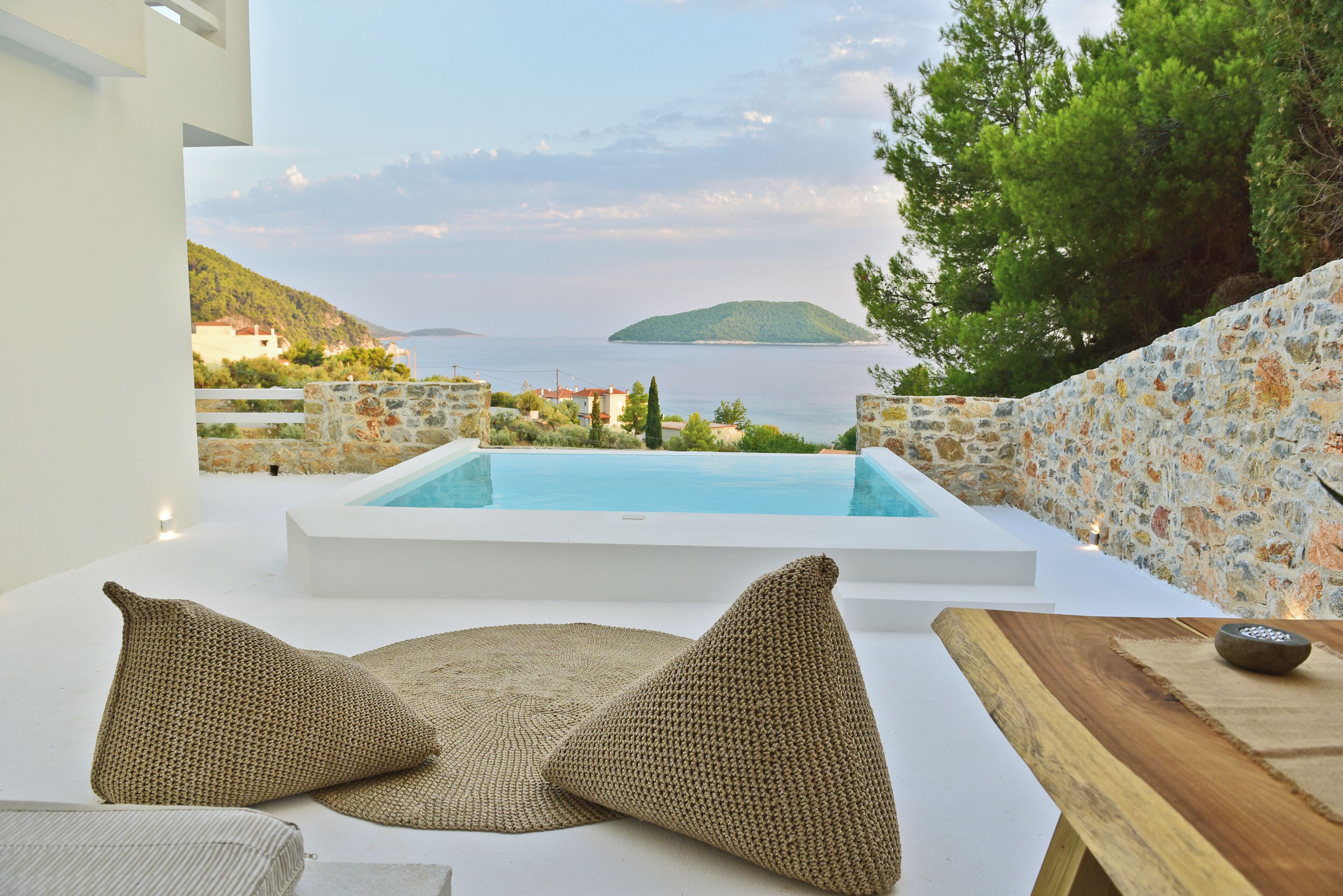 Zona junto a la piscina con vista al mar y colinas, una pared de piedra y una mesa de madera con un cuenco.