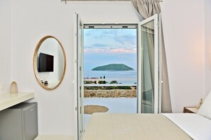 Une chambre avec un lit, une télévision, un petit réfrigérateur, un miroir et un balcon offrant une vue sur une île lointaine.