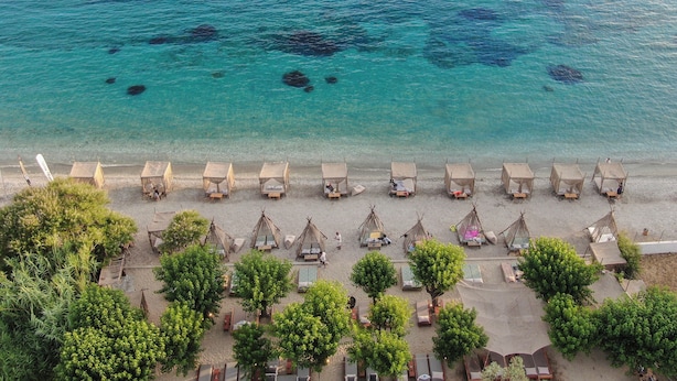 An aerial view of a beach with clear turquoise water, several thatched-roof huts, and lush green trees. - Natura Luxury Villas (Skopelos)