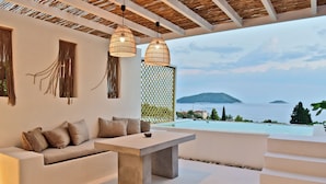 A modern outdoor living area with a concrete table, white sofa, and hanging wicker lights. - Natura Luxury Villas (Skopelos)