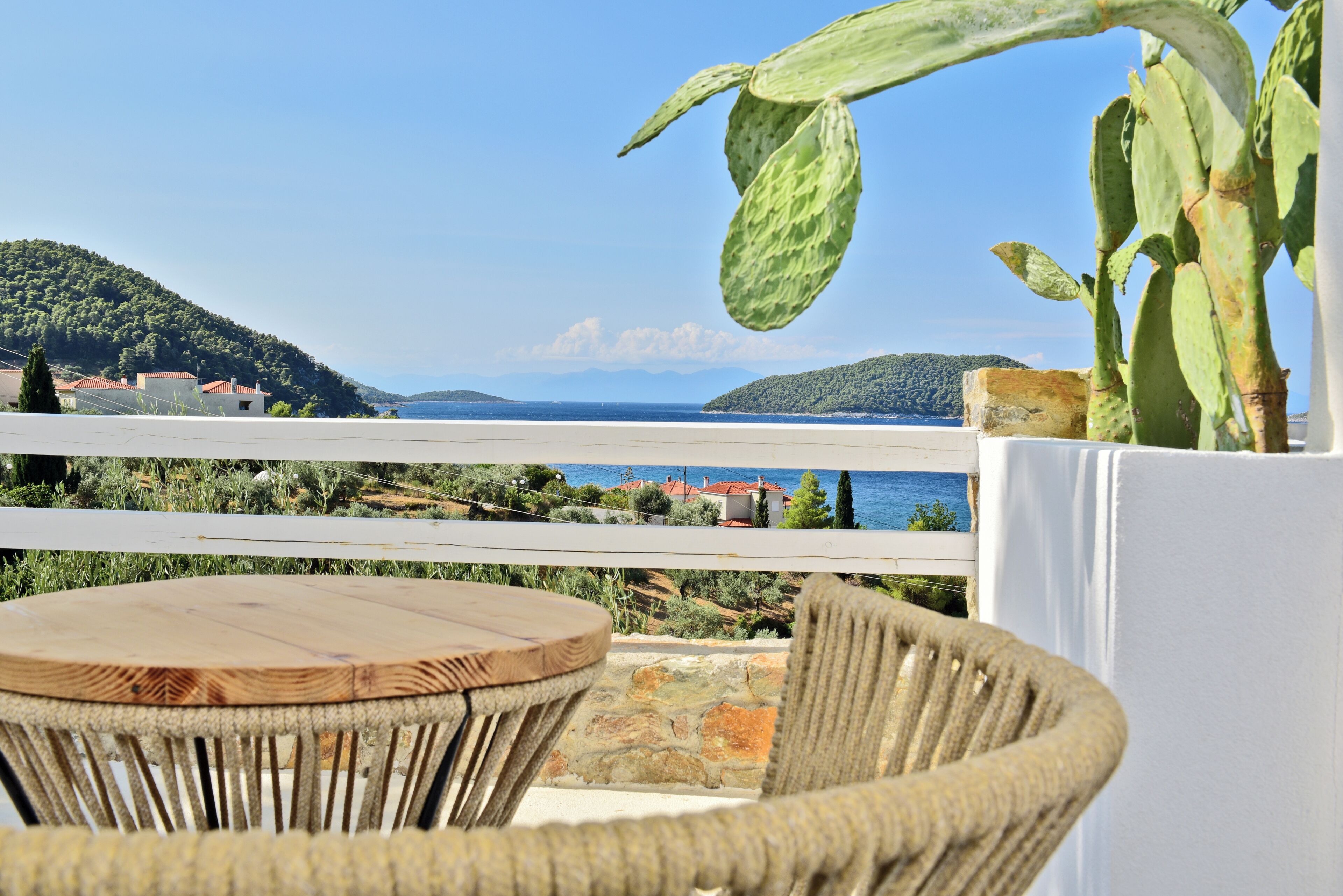 Una silla de mimbre y una mesa en un balcón con vistas a un paisaje costero que incluye el mar y colinas.