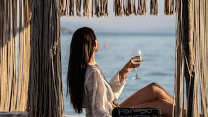 A person sitting under a woven structure by the sea, holding a glass. - Natura Luxury Villas (Skopelos)