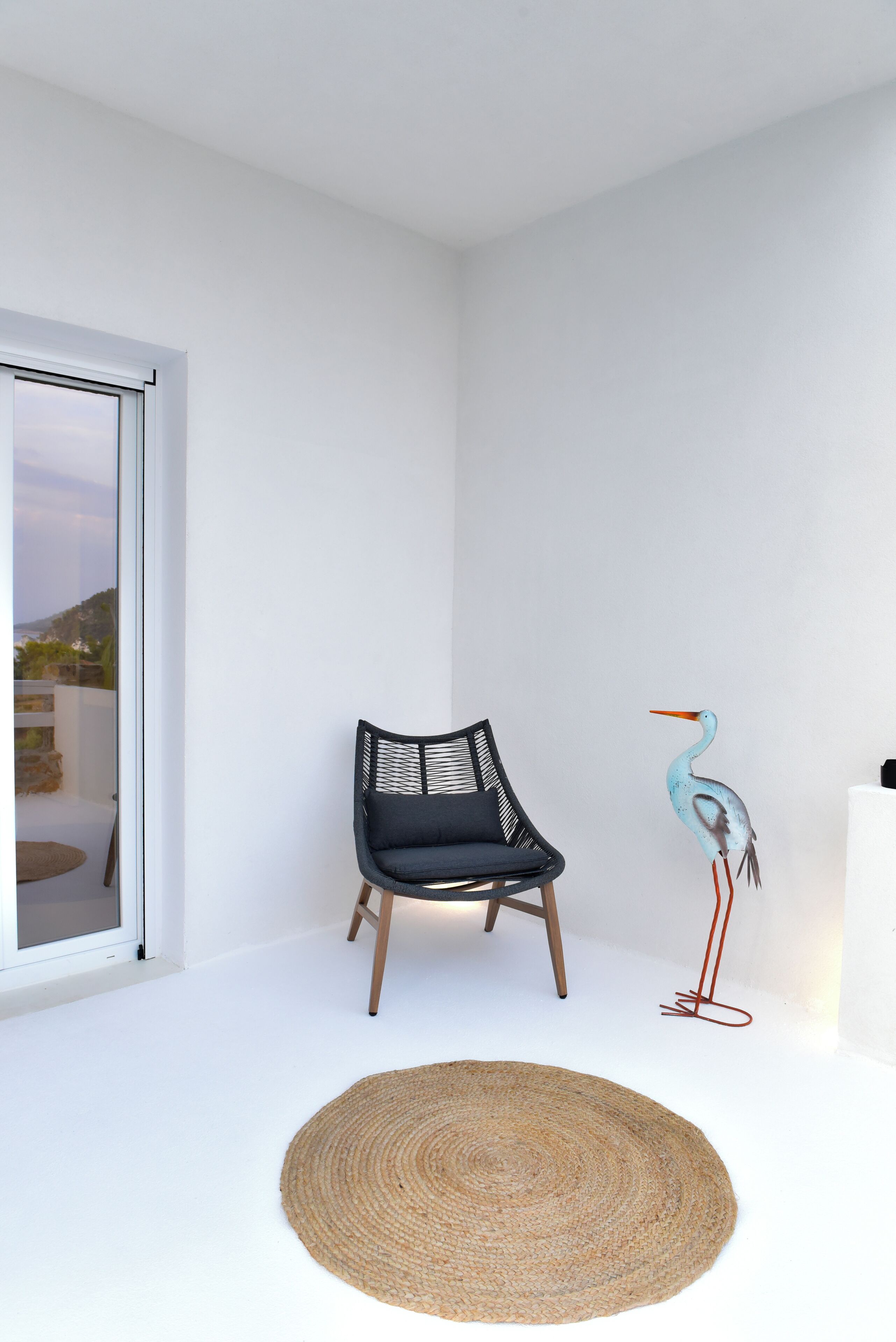 A modern room with a large window, a black chair with a woven backrest, a woven rug, and a decorative bird sculpture.