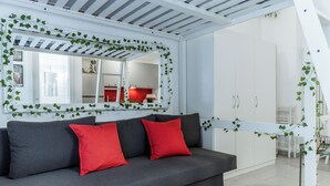 En moderne stue med en grå sofa, røde puder, et hvidt loft med en træbjælke og udsigt til et køkken og spiseområde i et stort spejl.