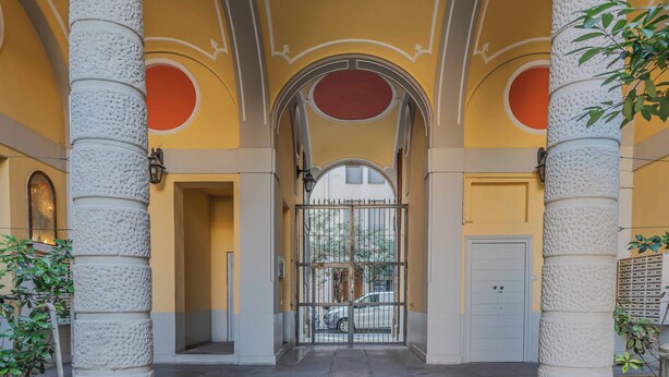 En imponerende indgang med en stor buegang, omgivet af høje søjler, der fører til en bygning med gul facade og røde detaljer.
