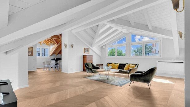 A spacious loft with high ceilings, exposed wooden beams, and large windows offering views of the outdoors.