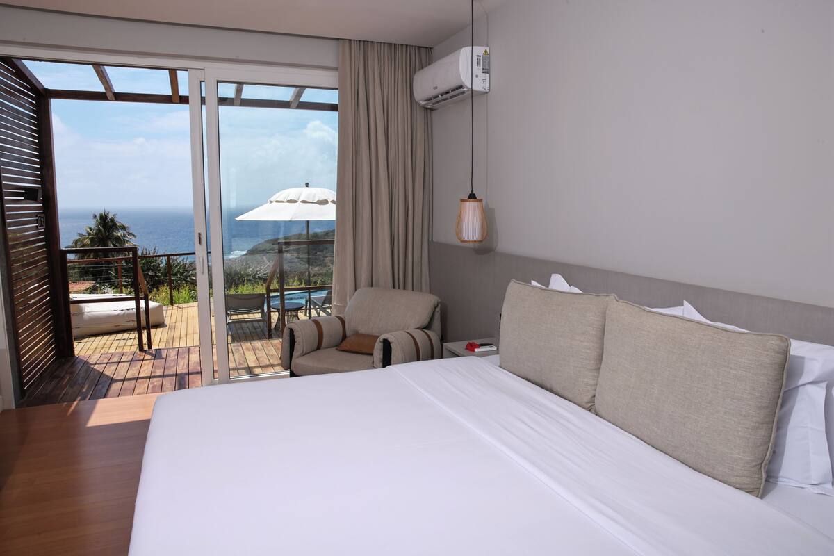 A hotel room with a large bed, a chair, a small table, and a view of the ocean through a sliding glass door.