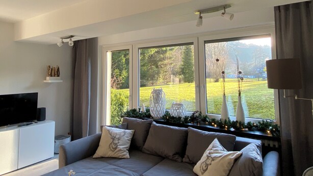 Ein Wohnzimmer mit einer Couch, einem Fernseher auf einer Kommode und einem großen Fenster mit Gartenblick.