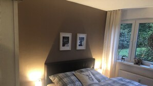 Ein Schlafzimmer mit einem Bett, zwei gerahmten Bildern an der Wand, einem Nachttisch mit Lampe und einem Fenster mit Blick ins Grüne.