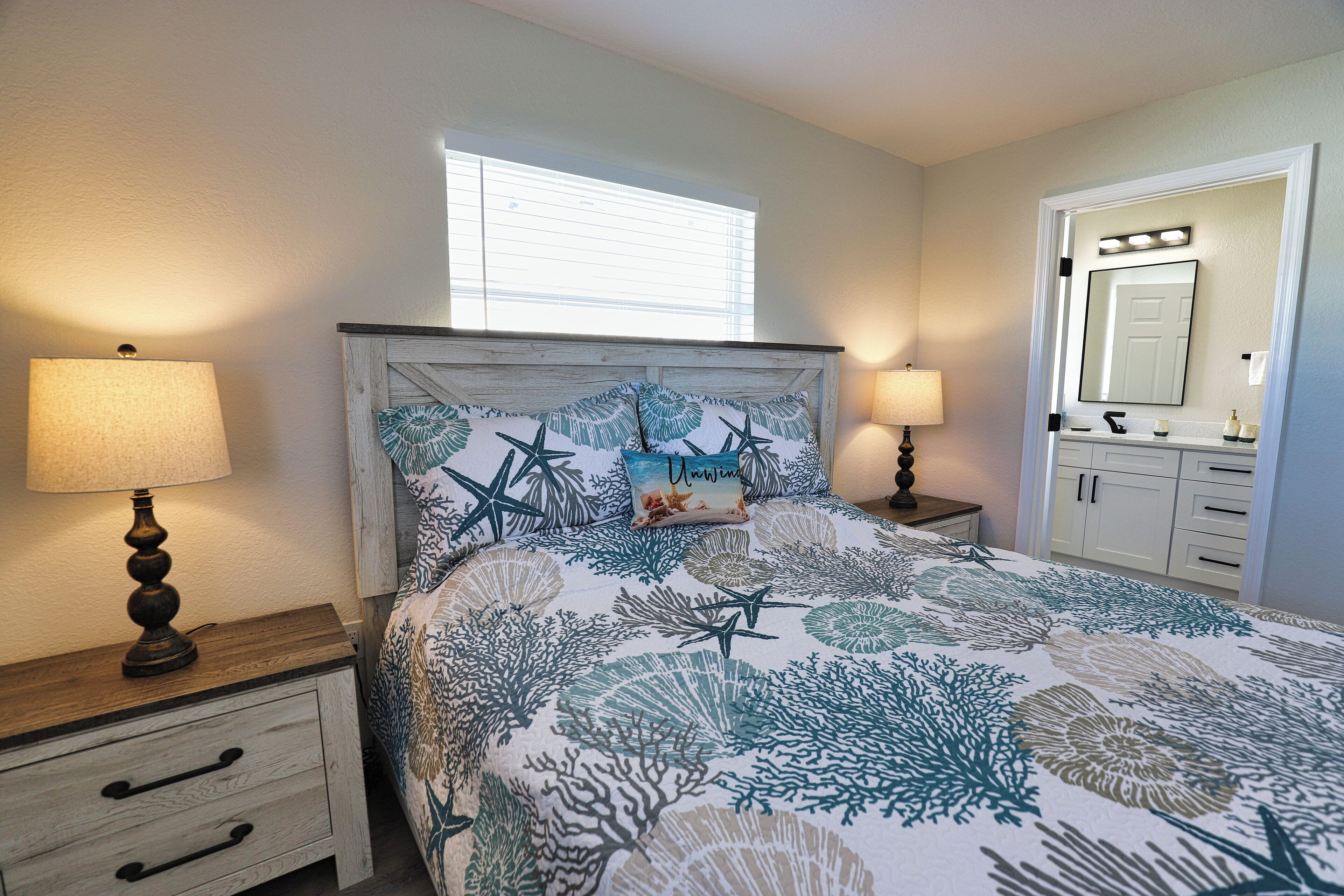 A bedroom with a bed, bedside lamps, a nightstand, a window, and a bathroom with a mirror and cabinets.