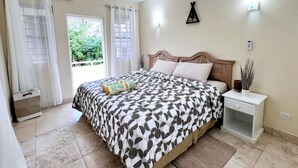 A bedroom with a bed, curtains, a bedside table, a nightstand, and a view of greenery outside. - Serene Barbados escape with ocean view - Peaceful (Christ Church)
