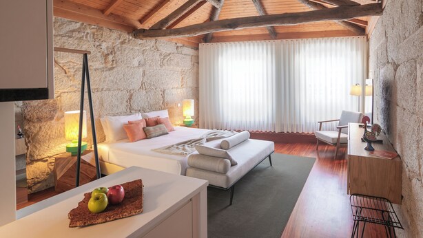 Una camera da letto con parete in pietra, travi in legno al soffitto, un letto con cuscini, un comodino con una lampada e una sedia.