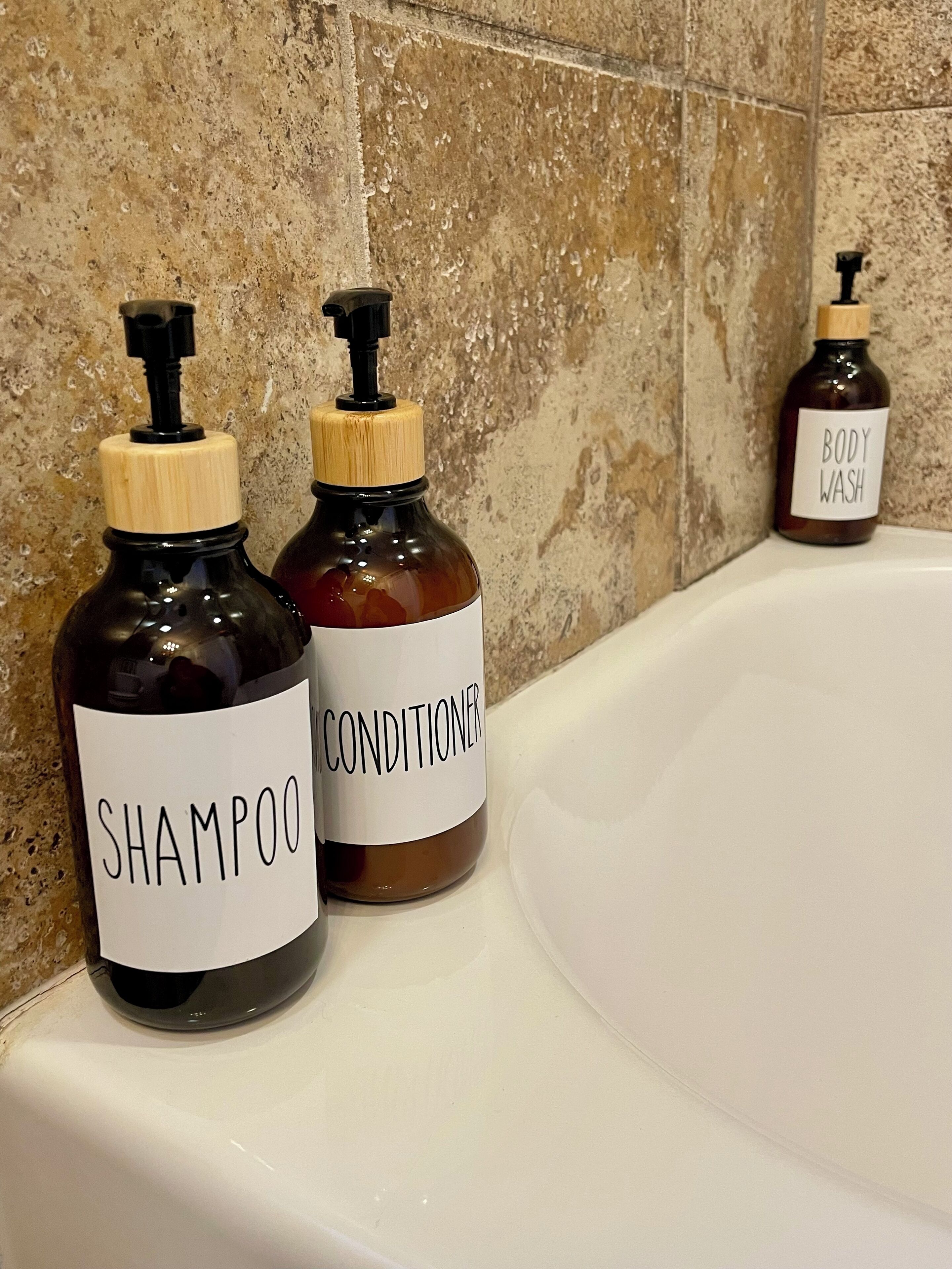 Kaksi pulloa shampoota ja hoitoainetta kylpyhuoneen tasolla, kylpyammeen vieressä.