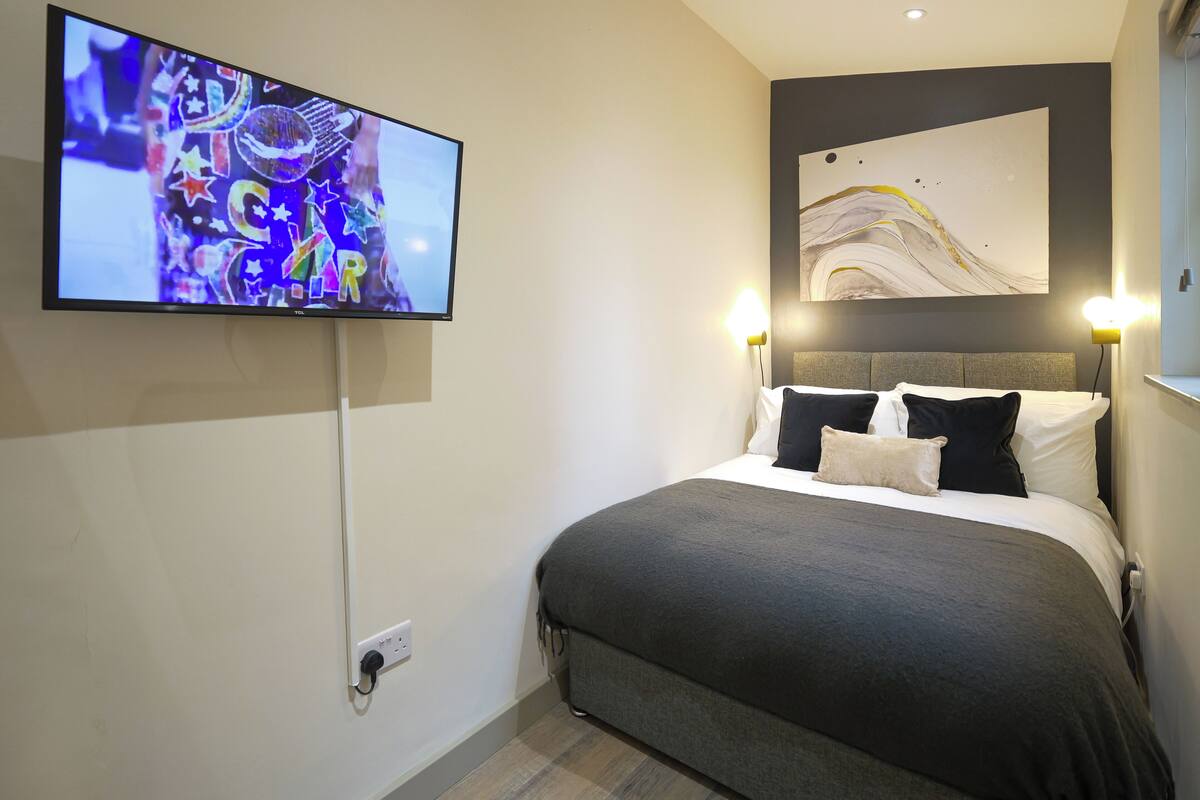 A modern hotel room with a large flat-screen TV mounted on the wall, a bed with grey bedding, two bedside lamps, and a large abstract painting above the bed.