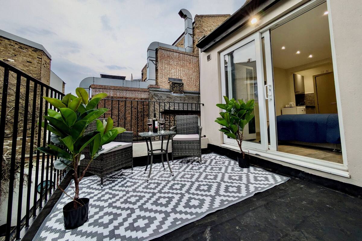A small patio area with a patterned rug, two potted plants, a table with chairs, and a glass door leading to a room with a bed and kitchenette.