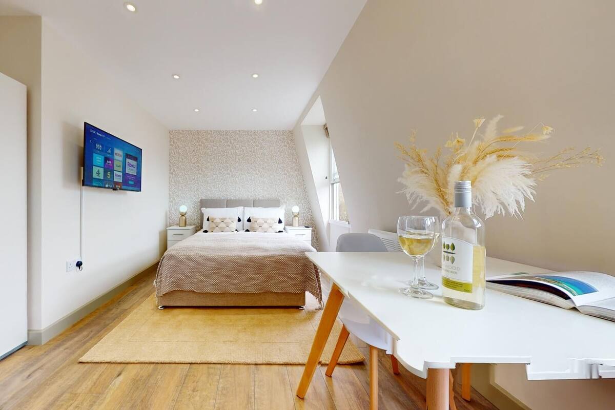 A modern bedroom with a bed, bedside tables, a TV, a dining table with a bottle and glass, and a window with natural light.