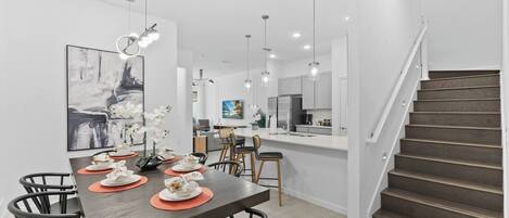 A modern kitchen with white cabinets, a dining area with a wooden table and chairs, and a staircase leading to an upper level.