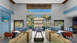 Eine geräumige Lobby mit einer zentralen Sitzgruppe, großen Fenstern mit Blick auf Palmen und einen Pool, und einem modernen architektonischen Design.
