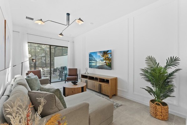 A modern living room with a sofa, armchairs, a coffee table, and a TV mounted on the wall. There is a large window with a view of the outdoors, and a potted plant on the right side. - Lux 5BR Home w/Game Room, 4 Suites & Private Pool (Orlando)