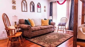 A living room with a brown sofa, two wooden chairs, a patterned rug, and a window with curtains. - Tinker AFB OKC I-40 Colonial Dorchester Cottage (Midwest City)