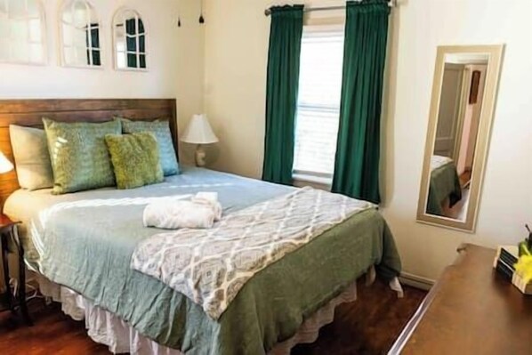 Une chambre avec une tête de lit en bois, un lit recouvert d’une couvre-lit à motifs, des rideaux verts et un miroir au mur.