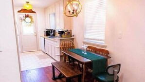 A kitchen with white cabinets, a wooden table with a green tablecloth, and a window with blinds. - Tinker AFB OKC I-40 Colonial Dorchester Cottage (Midwest City)