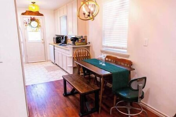 A kitchen with white cabinets, a wooden table with a green tablecloth, and a window with blinds. - Model City Homes Stays: Tinker AFB OKC I-40 Colonial Dorchester Cottage (Midwest City)