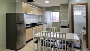 Een kleine keuken met een koelkast, fornuis en wastafel. Er is een eettafel met stoelen en een raam met gordijnen.