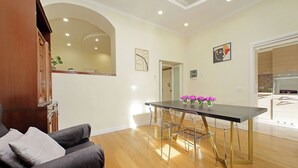 A modern living room with a dark sofa, a dining table with chairs, and a kitchen area visible through an archway.