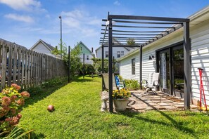 Un patio trasero residencial con cerco de madera, pérgola, patio de piedra y una casa de fachada blanca.