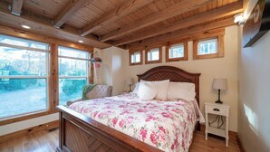 Ein Schlafzimmer mit einem Holzbettgestell, einer blumigen Bettdecke, einem Nachttisch mit Lampe und einem Fenster mit Blick auf Bäume.