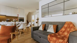Una sala moderna con un sofá gris, una manta color naranja y sillas blancas. Se ve una cocina con armarios blancos y un comedor con mesa y sillas al fondo.