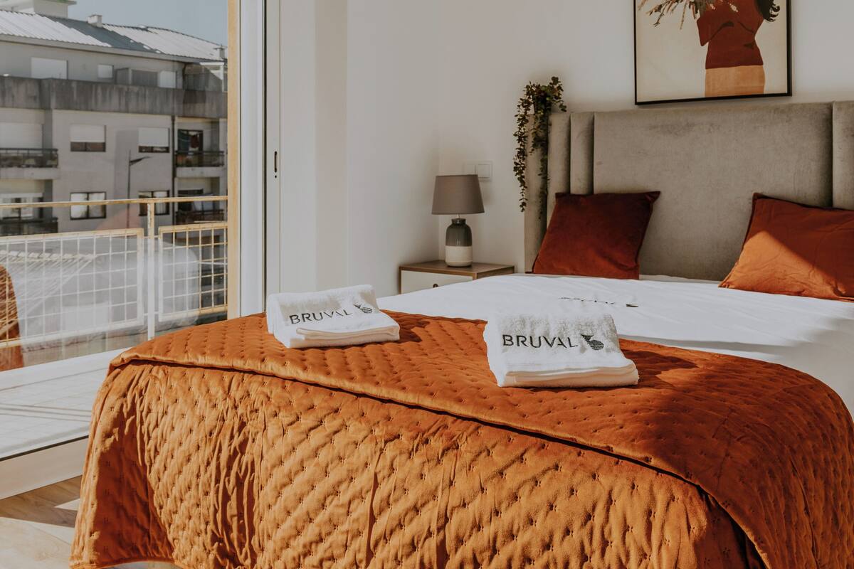 Ein modernes Hotelzimmer mit einem großen Bett, einem Nachttisch mit Lampe, einem gerahmten Kunstwerk an der Wand und Blick auf die Stadt durch das Fenster.