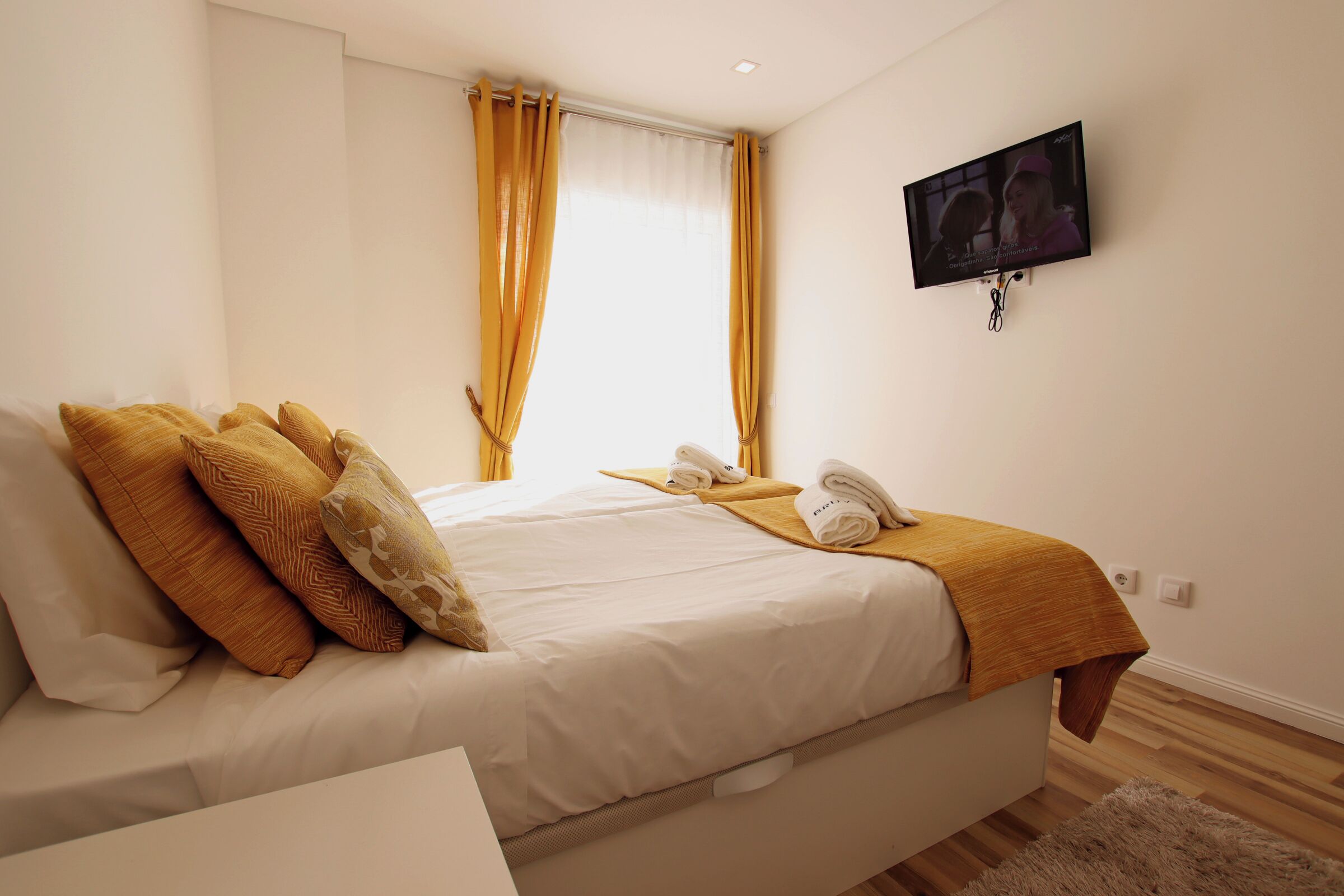 A neatly made bed with a white comforter and multiple pillows, a bedside table with a phone and a towel, and a window with yellow curtains.