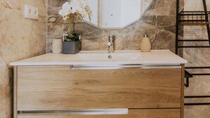 Un baño con pared de mármol, tocador de madera, espejo redondo y una orquídea en maceta.